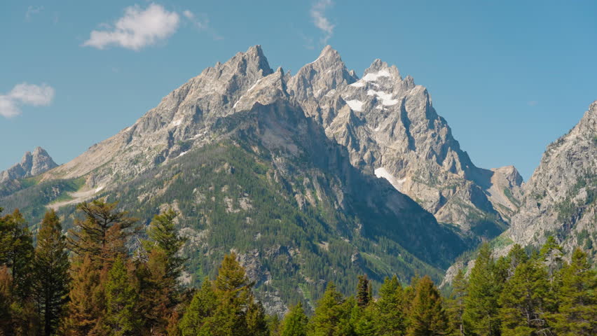 A towering mountain landscape rises dramatically above tall forest trees, creating a breathtaking contrast of rugged peaks and lush wilderness.