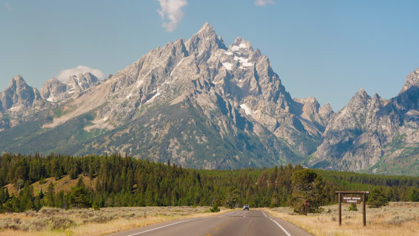 A scenic road stretches ahead, leading toward towering mountains and dense forest trees, creating a breathtaking journey through nature’s beauty.