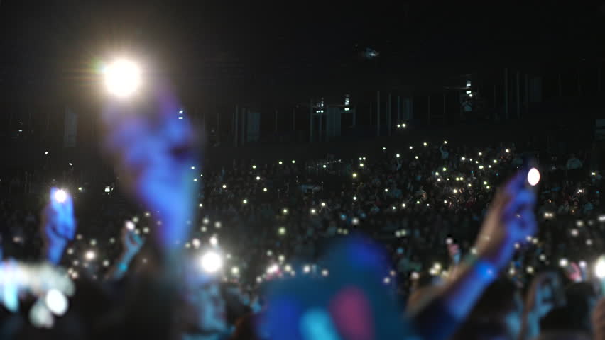 Many fun people lift hand up hold cell phone flash light. Fan crowd wave flashlights. Epic live music concert atmosphere. Big open air k pop arena. Cool night fest Lot joy men hang out. Kpop chill.