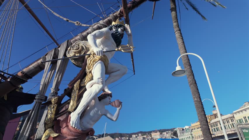 Close-up of a detailed Neptune figurehead on a historic ship in Genoa, Italy. The ornate sculpture with gold accents and maritime ropes contrasts against the clear blue sky and city buildings.