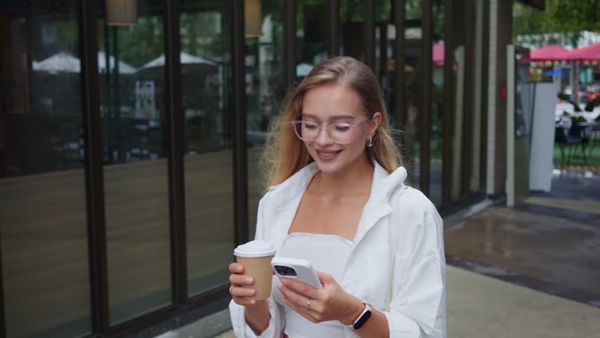 Young woman blogger enjoying coffee outdoors, blogger using smartphone and smiling brightly, blogger relaxing in stylish city setting, perfect footage for social media, lifestyle concepts.