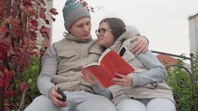 Romantic moment of lovers seated on park bench in autumn, woman reading book while man hugs her warmly holding phone, both dressed in cozy jackets, surrounded by red leaves and greenery - Powered by Shutterstock - Get 15% off with code: PIKWIZARD15