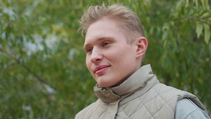 Young man kneels with warm genuine smile during emotional moment surrounded by swaying green leaves in soft breeze while wearing light quilted jacket