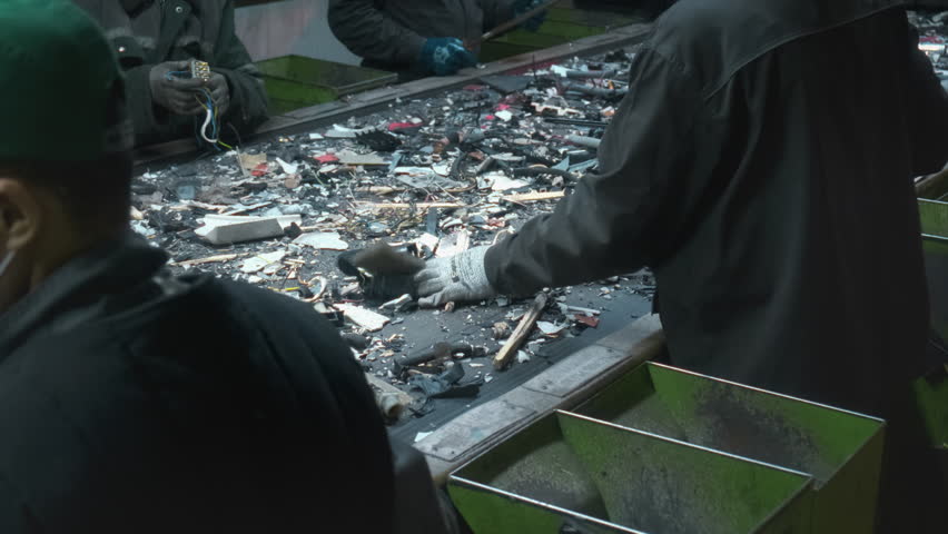 Sorting of waste on a conveyor belt. Male workers inspect a pile of garbage at a recycling facility. Unrecognizable men take care of the environment. Males touch trash made of iron and plastic