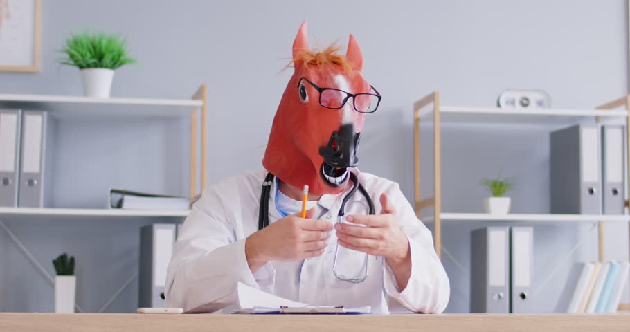 Portrait of unusual crazy man doctor or veterinarian wearing funny horse head mask and uniform sitting at office work desk with pencil paper giving professional online Internet consultation to patient