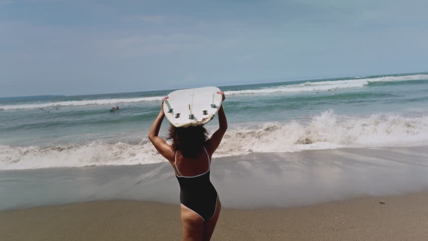 Happy old woman on beach holds a surfboard. Sporty elderly female on tropical island with sandy beach goes into water, enters the sea or ocean. Bali coast. Wave for surfing. Senior retired woman