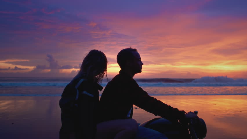 Romantic couple on motorcycle rides along a sandy black beach in Bali. Man and woman at sunset drive bike. Female and male make an adventure, road trip. Twilight and the blue hour.