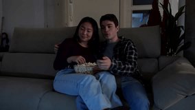 Multiethnic couple relaxing on sofa, watching television, using remote control, and eating popcorn - Powered by Shutterstock - Get 15% off with code: PIKWIZARD15