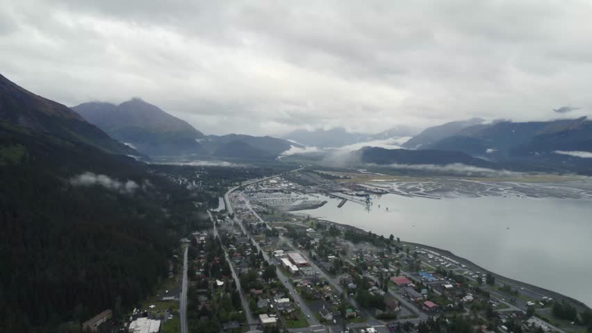 Aerial establishing shot Seward Town in Alaska, USA. CLoudy day in residential area with mountain range in background. Port and pier in distance. Wide shot.