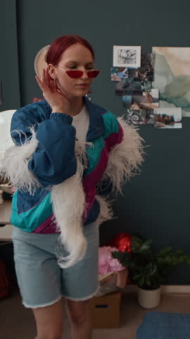 Vertical view of hands of unrecognisable girl shooting smartphone video of cheerful red-haired female friend posing with white boa at home