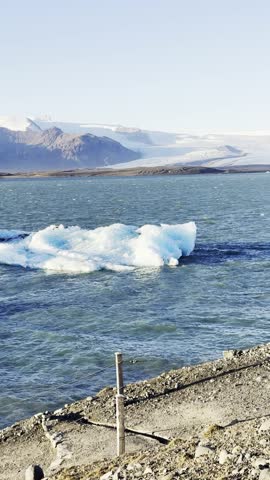 A vertical view of shore with icebergs on the water on a sunny day, showcasing people on the beach and ice-covered mountain in the backdrop in Iceland