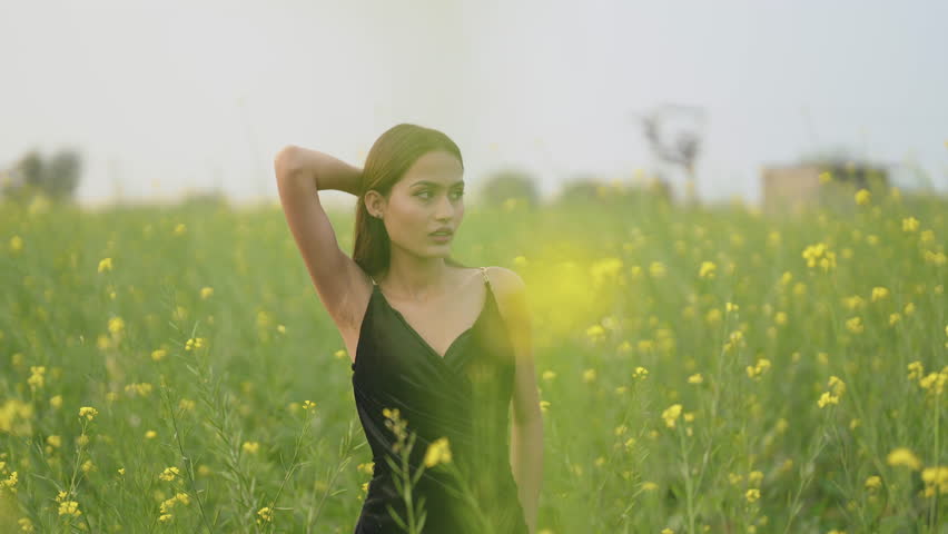 Indian happy young adult gen z beautiful model woman wear black dress walking outdoor enjoy nature. Asian smiling relax teen age girl looking at camera touching yellow flower outside