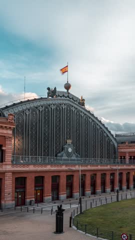 Majestic atocha train station, a vital transportation hub in madrid, stands proudly beneath the waving spanish flag.timelapse view.