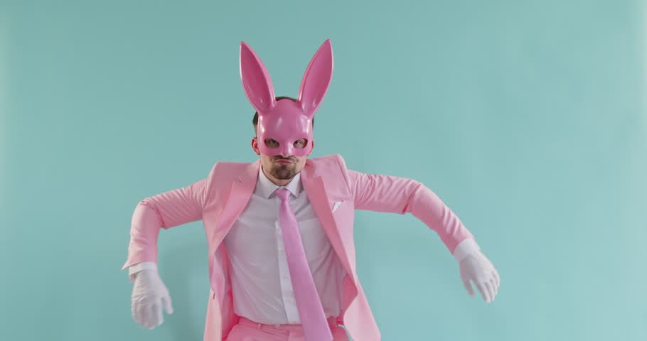 A man in a pink suit and a bunny mask dances merrily against a blue background. Concept of a funny easter dancer in a mask. Bright elegant bunny costume for a party. Modern corporate entertainment