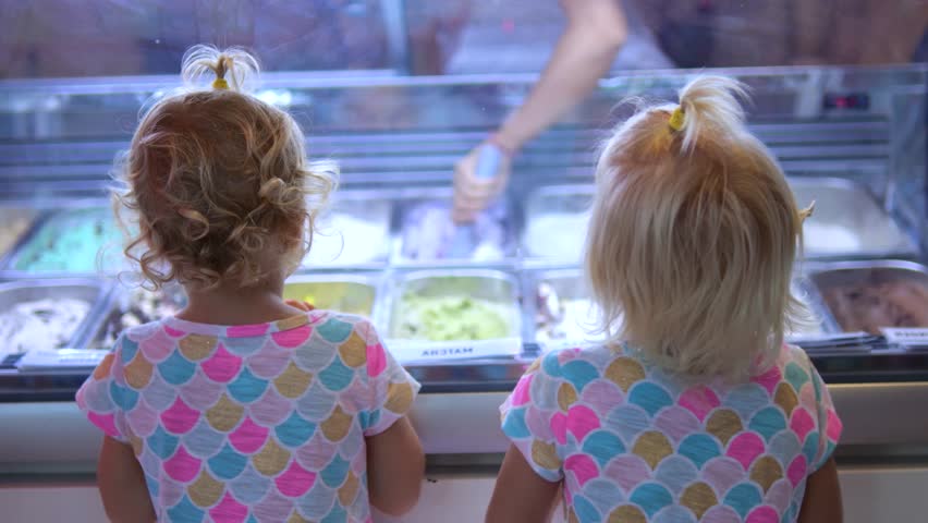 Two little girls with similar hairstyle and shirts are standing in front of an ice cream display, looking at the various flavors and trying to decide what to choose