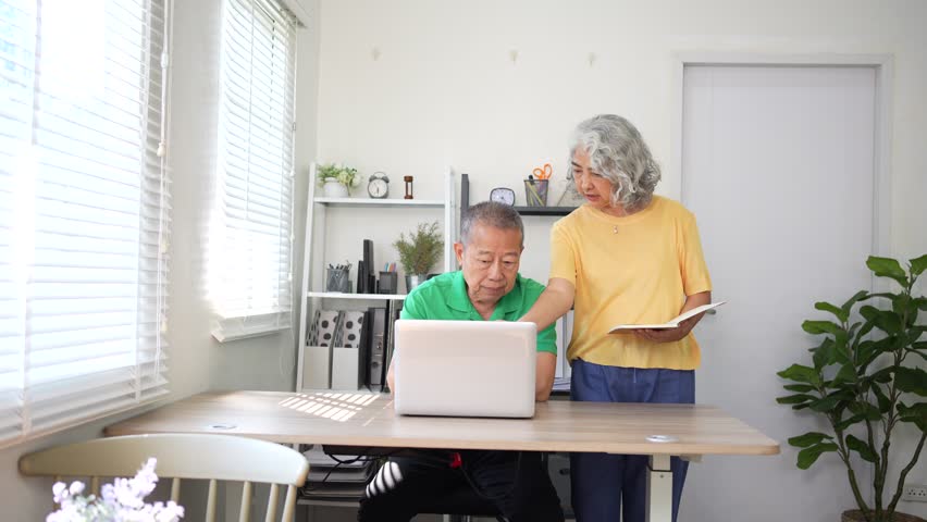 Elderly couple working together at home with teamwork, man using laptop while woman holds clipboard and reviews notes.