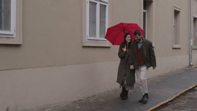 Romantic young couple walking closely under bright red umbrella, sharing tender moments while strolling through rainy urban street during autumn season - Powered by Shutterstock - Get 15% off with code: PIKWIZARD15