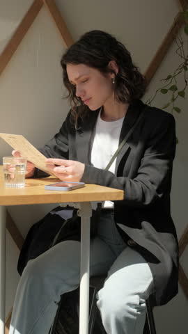 Сharming curly brunette woman sitting in cozy cafe, reading menu and choosing breakfast, brunch, or dessert with coffee. elegant female customer enjoying relaxing atmosphere in a modern coffee shop