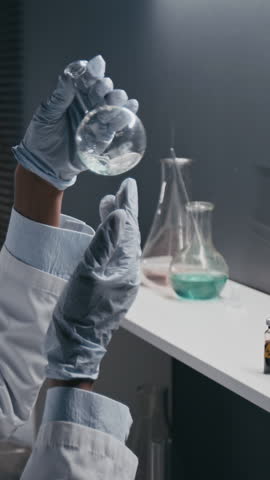 Vertical side shot of anonymous scientist in protective gloves holding flask with solution and adding reagent, working in drug development laboratory