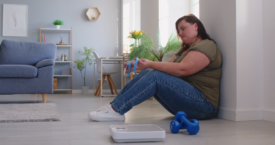 Upset overweight woman sitting on floor at home by scales and dumbbells, feeling distressed. Plus size female with measuring tape showing body insecurity, health concerns and problem with weight loss.