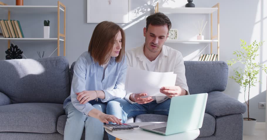 Focused confused young family married couple at laptop, reading financial papers, counting troubled with high expenses, life costs, worried spouses and debt, bankruptcy, small family business loss