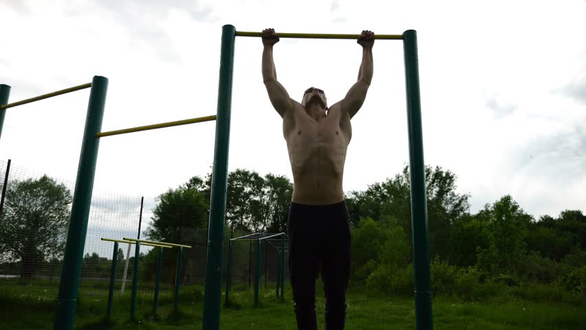 Muscular athlete doing pull ups on horizontal bar at sports ground. Young shirtless sportsman exercising at nature. Strong sporty man training outdoor. Concept of healthy and active lifestyle. Slow mo