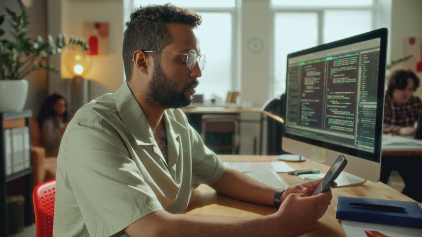 Concentrated male software developer sitting at desk in modern open plan office, checking smartphone as code runs on computer monitor, colleagues working in background
