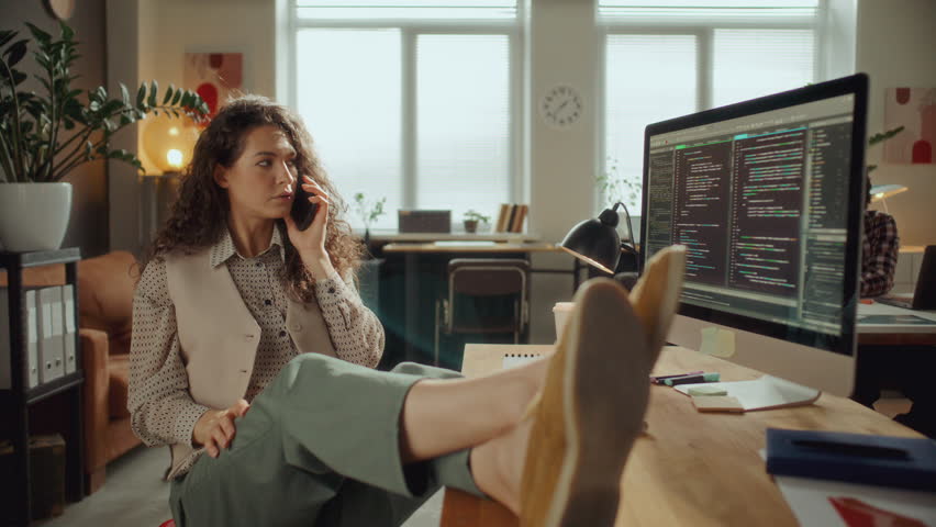 Young female software developer sitting comfortably with feet on desk, chatting on cell phone and smiling during workday in open plan office, programming code displayed on computer screen
