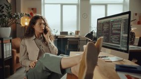 Young female software developer sitting comfortably with feet on desk, chatting on cell phone and smiling during workday in open plan office, programming code displayed on computer screen - Powered by Shutterstock - Get 15% off with code: PIKWIZARD15