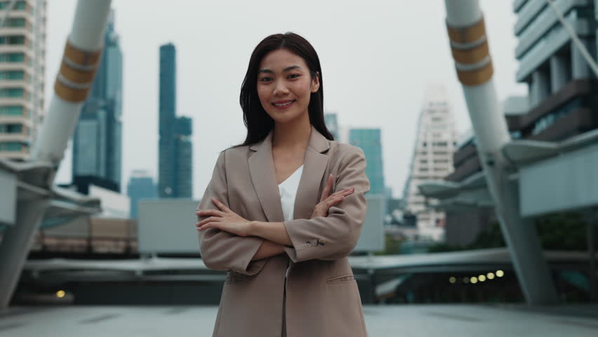 Portrait of young Asian female with confident smile crossing arms standing in megapolis environment. Thai business lady shows determination to reach success. Stylish corporate worker