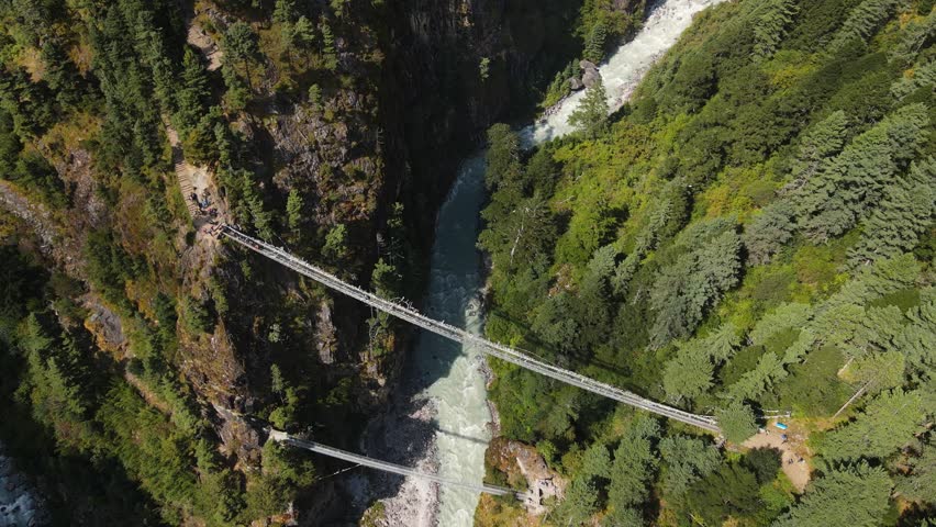 Two suspension bridges, one above the other, span a deep gorge in Nepal. The river below cuts through steep cliffs, surrounded by dense pine forests and rugged mountain terrain.