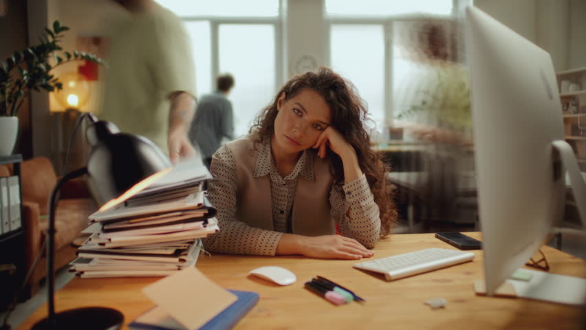 Portrait of young frustrated woman resting head on hand and looking into camera while sitting at desk in open plan office, overloaded with paperwork stacked nearby. Time lapse