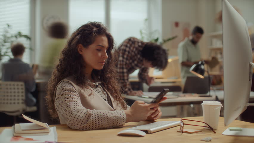 Young beautiful woman with curly hair sitting at desk in modern open space office, typing on smartphone and smiling, while coworkers moving around in background. Time lapse