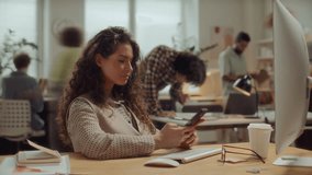 Young beautiful woman with curly hair sitting at desk in modern open space office, typing on smartphone and smiling, while coworkers moving around in background. Time lapse - Powered by Shutterstock - Get 15% off with code: PIKWIZARD15