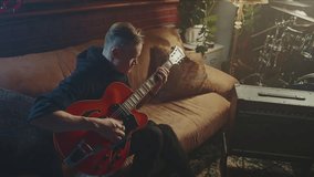 Professional musician composing original melody, playing red electric guitar while sitting comfortably on sofa in home recording studio, capturing creative musical moment with soft ambient lighting - Powered by Shutterstock - Get 15% off with code: PIKWIZARD15