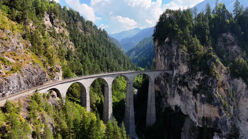 Drone footage of a train emerging from a tunnel and crossing the iconic Landwasser Viaduct in the Swiss Alps