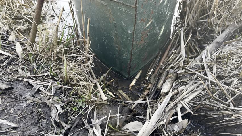 old fishing boat, rusty metal, stands tied up among dry reeds on the bank of River, pond or lake with Rocky shore
