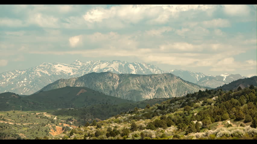 Timelapse mountains. Mountain ranges, green slope in foreground. Cloudy sky cumulus clouds quickly run across blue sky. Snow-capped mountain peaks are preserved in distance. Cinematic video background
