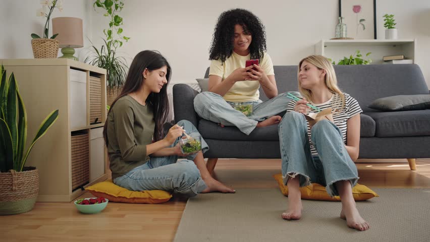 Flatmate girl friends having fun looking at cell phone app while laughing and enjoying breakfast together at home