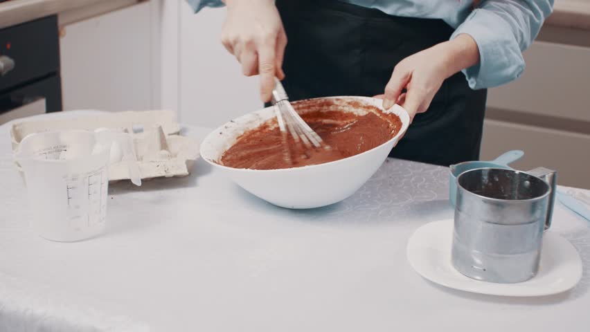 A woman makes chocolate dough. A female whisks dough at home in the kitchen. A housewife makes cupcakes.