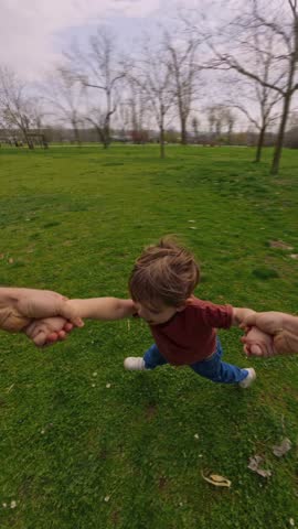 Parent holds the hands of their happy laughing child and spins them around in a circle in the park. POV view, father playing with his joyful son outdoors. Happy childhood concept. Vertical video