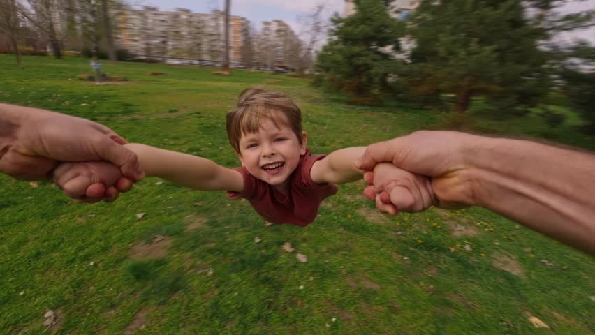 First-person view, father holding his son's hands, spinning him around in a circle at the park, joyful child laughs, enjoying the playful moment, creating happy childhood and family memories together