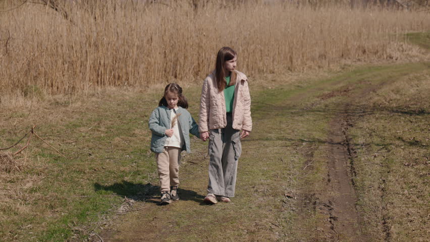 Two sisters walk hand in hand along a grassy country path, enjoying a quiet spring day surrounded by tall dry reeds and warm sunlight. High quality 4k footage