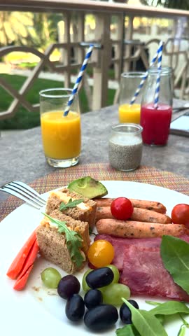 Breakfast plate with sausages, avocado, grapes, bread, and cold cuts on outdoor table, fresh sausages grilled perfectly, nutritious sausages offer healthy start, delicious dining scene.