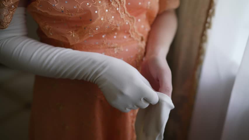 A vintage-style woman carefully puts on thin white long gloves, embodying the sophisticated glamour of the 1920s through precise, elegant hand movements near the soft light from the window.