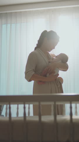 Young mother standing near large window, holding her newborn baby in arms and gently stroking her in slow-motion