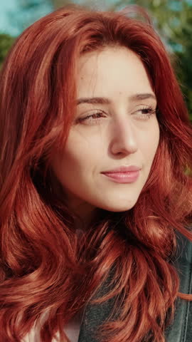 Vertical video, Beautiful redhead woman smiling and looking to the side in a park. She is wearing a dark jacket and has long, flowing red hair.