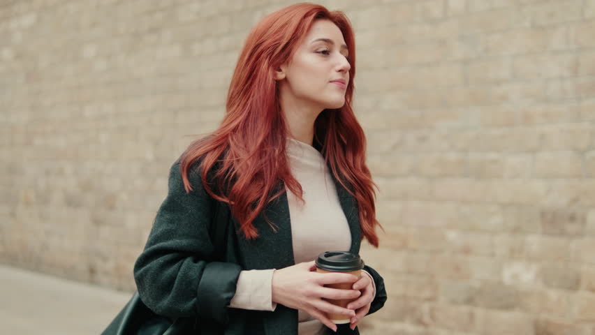 A young woman with long, red hair walks down a city street while enjoying her coffee. She takes in the sights and sounds of urban life during a peaceful stroll.