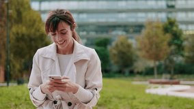 A cheerful young lady is using her phone in a park with an office building in the background. She smiles and types a message. - Powered by Shutterstock - Get 15% off with code: PIKWIZARD15