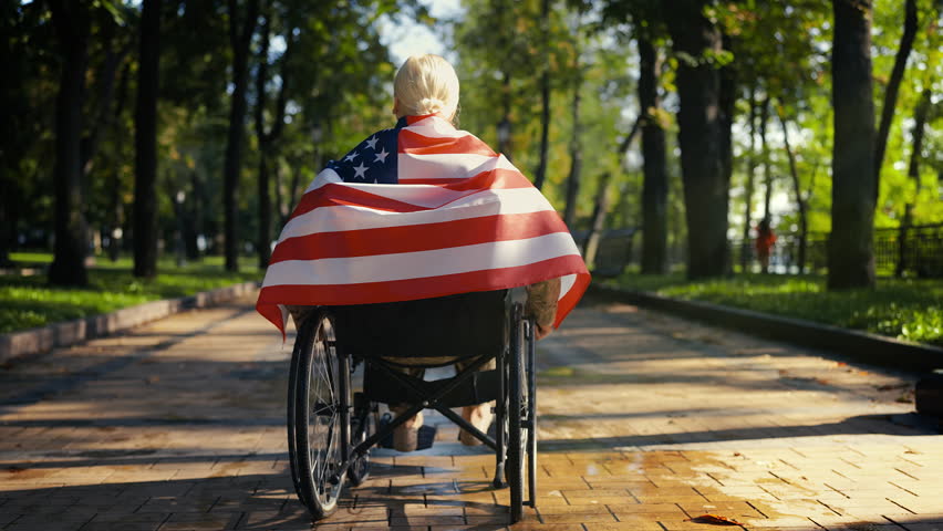 Woman army veteran rides a wheelchair wrapped in US flag, proud and reflective